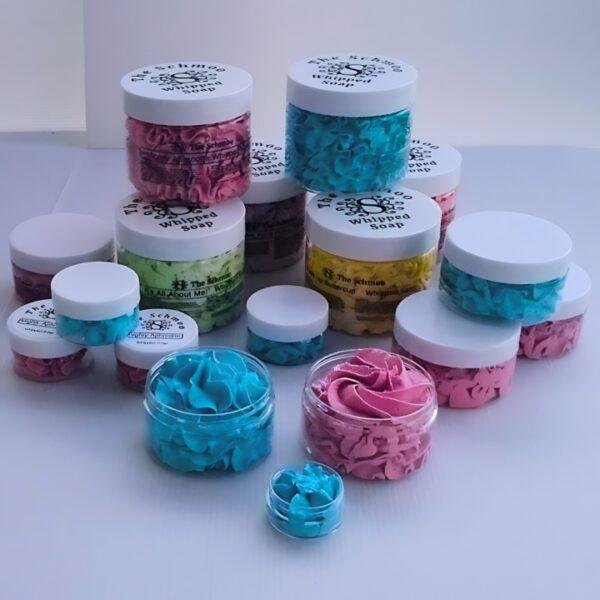 A collection of The Schmoo’s Whipped Soaps in assorted bright colours and fragrances, displayed in jars of different sizes.